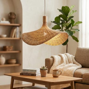 Peut inclure: Suspension luminaire en rotin tressé avec une partie supérieure en bois et un design incurvé et ouvert. La lampe est suspendue au-dessus d'une table basse en bois avec des livres, une plante en pot et une tasse. La pièce a une palette de couleurs neutres.