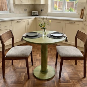 May include: A small, round, green table with a single pedestal base, set for two. Two wooden chairs with grey upholstered seats are positioned on either side. The table is set with plates and placemats. A vase of flowers sits in the centre.