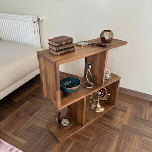 Puede incluir: Una mesa auxiliar de madera con un diseño geométrico único. La mesa está llena de objetos decorativos, incluyendo libros, una lupa, un cuenco, un candelabro y una brújula. La mesa es de madera de tonos cálidos.