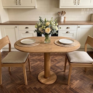 Op de afbeelding: Een ronde houten eettafel met twee bijpassende stoelen. De tafel heeft een enkele voet en is gedekt met twee borden en placemats. Een vaas met bloemen staat in het midden. De stoelen hebben beige kussens. Tafeldiameter ca. 90 cm.