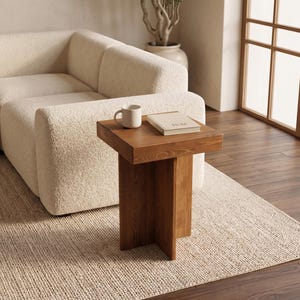 May include: A square, wooden side table with a natural finish. A white mug and a book rest on the table. The table is next to a cream-colored sectional sofa and a woven rug. A decorative plant is in the background.
