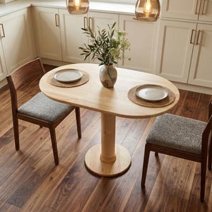 May include: A small, light wood oval dining table with two matching chairs. The table has a single pedestal base and is set with two plates on woven placemats. A vase of greenery sits in the centre. The chairs have grey upholstered seats.