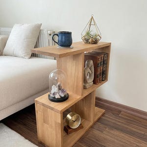 Puede incluir: Una mesa auxiliar de madera clara con un diseño abierto único. La mesa tiene varios estantes que sostienen una taza azul, un terrario, libros, una escultura, un reloj y una campana de cristal con cristales. La mesa está junto a un sofá beige.