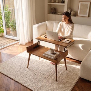 May include: A two-tiered, wooden lift-top coffee table with a laptop on the top tier. The table is brown with angled legs and a lower shelf. A white rug and a white sofa are in the background.