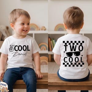 May include: A white short-sleeve t-shirt featuring a black graphic on the front with a peace sign, the text "COOL" and the name "Daniel". The back of the shirt displays a black checkered pattern, the text "TWO COOL" and a pair of sunglasses.