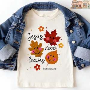 May include: A cream-colored short-sleeve t-shirt layered under a blue denim jacket. The shirt features a graphic with three smiling autumn leaves in red, orange, and brown, with the text "Jesus never leaves" and "Deuteronomy 31:8" in black.