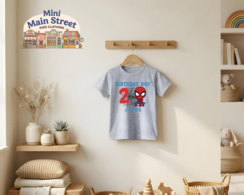 May include: A gray t-shirt with a Spider-Man graphic, the number 2, and the name "James". The shirt says "Birthday Boy" in blue text. The shirt is hanging on a wooden hanger against a neutral wall.