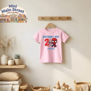 May include: A pink t-shirt featuring a Spider-Man graphic with the text "BIRTHDAY BOY" and the number "2". The name "JAMES" is printed below the graphic. The shirt is hanging on a wooden hanger.