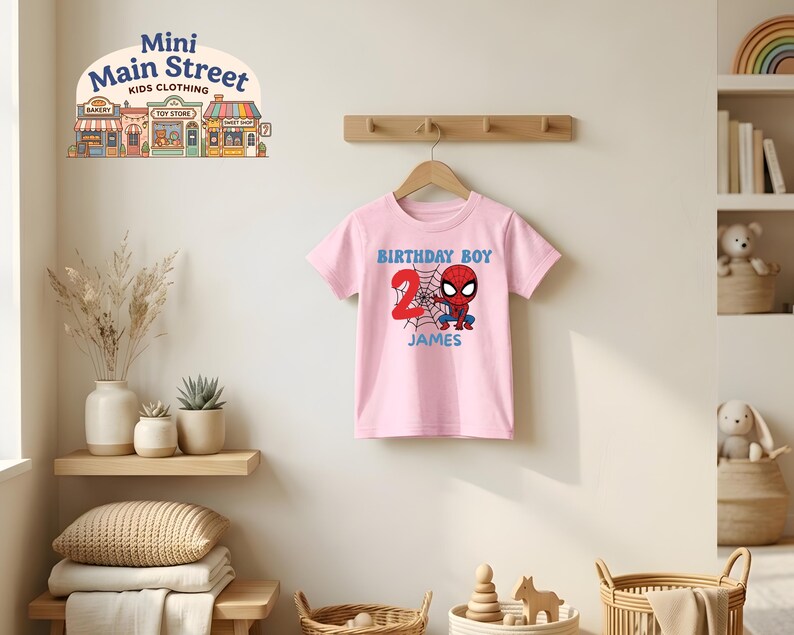 May include: A pink t-shirt featuring a Spider-Man graphic with the text "BIRTHDAY BOY" and the number "2". The name "JAMES" is printed below the graphic. The shirt is hanging on a wooden hanger.