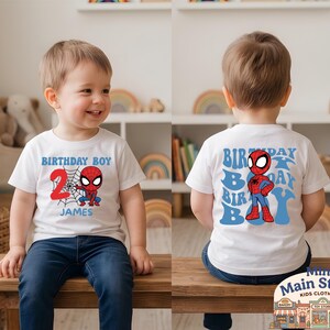 May include: White t-shirt featuring a Spider-Man graphic for a 2nd birthday. The front displays "BIRTHDAY BOY", the number "2", a web, and the name "JAMES". The back has a Spider-Man graphic and "BIRTHDAY BOY" in blue.