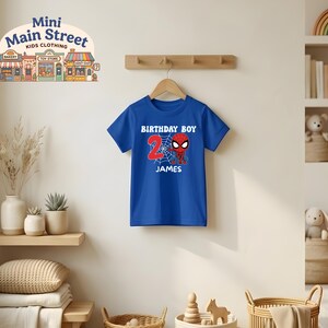 May include: A blue t-shirt with a Spider-Man graphic for a 2-year-old's birthday. The shirt features the words "BIRTHDAY BOY", the number "2", the name "JAMES", and a Spider-Man design. The shirt is hanging on a wooden hanger.