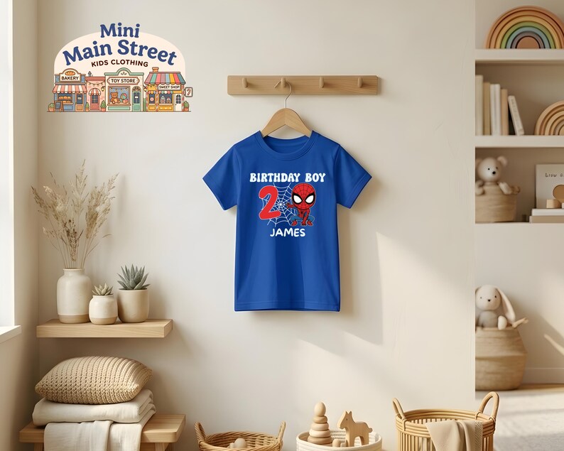 May include: A blue t-shirt with a Spider-Man graphic for a 2-year-old's birthday. The shirt features the words "BIRTHDAY BOY", the number "2", the name "JAMES", and a Spider-Man design. The shirt is hanging on a wooden hanger.