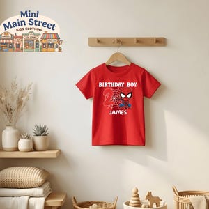 May include: A red t-shirt with a Spider-Man graphic, the words "BIRTHDAY BOY", the number "2", and the name "JAMES" in white. The shirt is hanging on a wooden hanger against a white wall. The image includes a "Mini Main Street Kids Clothing" logo.