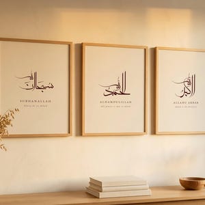 Puede incluir: Tres impresiones artísticas enmarcadas con caligrafía árabe en una habitación de tonos neutros. Cada impresión tiene un marco de madera y presenta una escritura elegante con traducciones en inglés debajo. La habitación está decorada con un jarrón de plantas secas y una mesa de madera.