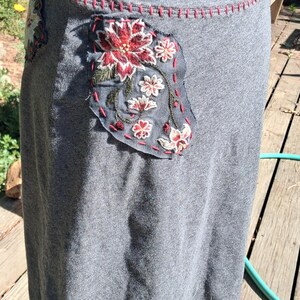 May include: A heather gray skirt featuring a wide waistband and a hemline both accented with thick red decorative stitching. The skirt includes an appliqued patch on the front displaying intricate red and white floral embroidery on dark fabric.