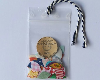 Può includere: Un sacchetto di plastica trasparente contenente pezzi di tessuto colorati in vari modelli e colori. Un'etichetta rotonda con le parole "HAPPY ANNIVERSARY" e "TAILORBIRD DESIGNS" è attaccata. Il sacchetto è legato con un cordoncino intrecciato blu e bianco.