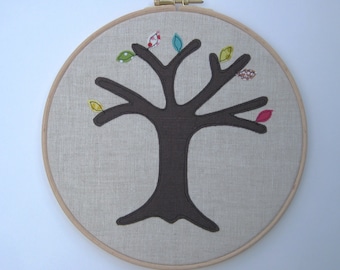 Può includere: Un albero di stoffa marrone con foglie di stoffa colorate è cucito su uno sfondo di lino beige e incorniciato in un cerchio di legno.