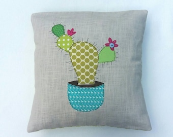 Può includere: Un cuscino grigio con un cactus verde e giallo con fiori rosa in un vaso turchese con un motivo bianco. Il cactus è fatto di tessuto e ha un design cucito.