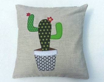Può includere: Un cuscino beige con un'applicazione di cactus verde. Il cactus ha fiori rossi ed è in un vaso con motivo bianco e nero.
