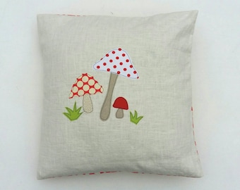 Può includere: Un cuscino beige con bordo a pois rossi e bianchi presenta un'applicazione rossa, bianca e verde di tre funghi.