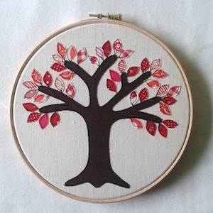 May include: A handmade tree design on canvas, framed in a wooden embroidery hoop. The tree has a dark brown trunk and branches, decorated with red and pink fabric leaves. The leaves display diverse patterns, including polka dots and stripes.