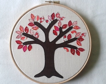 Può includere: Un design ad albero fatto a mano su tela, teso in un telaio da ricamo in legno. L'albero presenta un tronco e rami marrone scuro, adornati da foglie di tessuto rosse e rosa. Le foglie hanno vari motivi, tra cui pois e strisce.