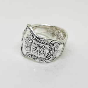 Puede incluir: Un anillo de plata con un diseño detallado que presenta un escudo con un león y otros símbolos. El anillo está hecho de una cuchara reutilizada.