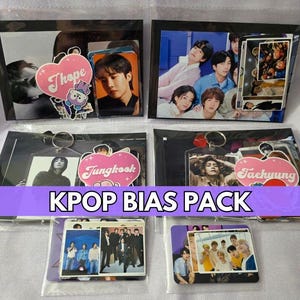 May include: Kpop Bias Pack featuring photo cards, keychains, and stickers. Each pack includes images of K-pop stars. The packs are in clear plastic sleeves with black backing. The text "KPOP BIAS PACK" is displayed in a purple banner.