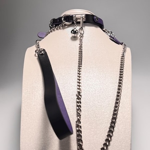Puede incluir: Un collar negro y morado con una cadena y una campana plateadas. El collar se muestra en una cabeza de maniquí blanca. Una paleta negra con un interior morado está unida al collar.