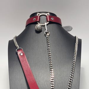 Puede incluir: Un collar de cuero burdeos con un anillo plateado en forma de gato y una bola plateada colgante. Se adjuntan una cadena plateada y una correa de cuero burdeos. El collar se exhibe en un maniquí negro.