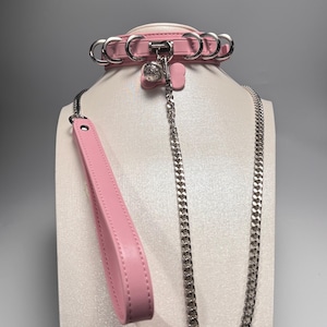 May include: A pink leather pet collar with a matching leash and a silver chain. The collar has silver rings and a small charm. The leash has a handle. The set is displayed on a white mannequin.