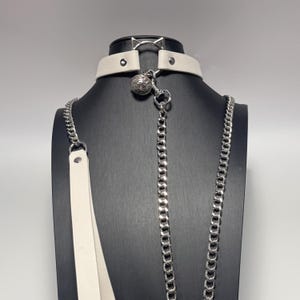 Puede incluir: Un collar de cuero blanco con una cadena de metal plateado y un colgante de bola decorativa. El collar tiene un anillo de metal plateado y se muestra en un maniquí negro. La cadena se extiende hacia abajo, sugiriendo una correa.