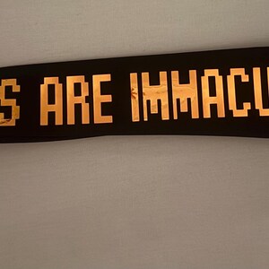 May include: Black necktie with gold text that reads "VIBES ARE IMMACULATE HERE." The tie is made of a dark material and features a standard tie knot.