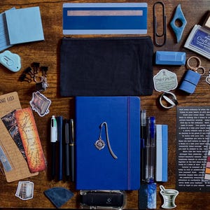 May include: A collection of stationery items, including a blue journal with a compass bookmark, pens, sticky notes, a black pencil case, and a stamp pad. The items are arranged on a wooden surface, creating a visually appealing collection.