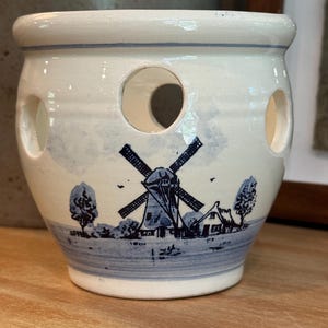 May include: A cream-colored ceramic pot featuring a blue painted landscape of a windmill and small houses. The vessel has a wide rim and several circular cutouts around the upper body, with a blue band painted near the base and the top rim.