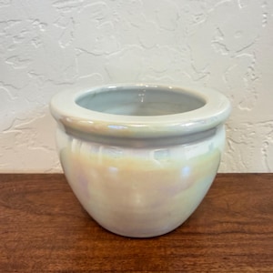 May include: A small, round ceramic planter featuring a smooth, iridescent glaze that reflects soft hues of yellow, pink, and blue. The bowl-shaped vessel has a thick, rounded rim and a wide opening, with a glossy finish that catches the light.