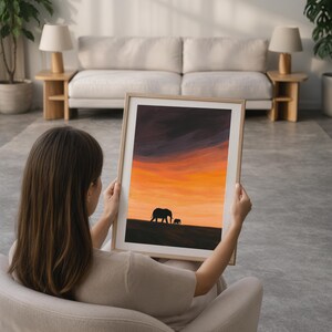 May include: A framed art print featuring a sunset scene with silhouettes of two elephants. The sky transitions from dark purple to vibrant orange and yellow. The artwork is displayed in a light-colored wooden frame.