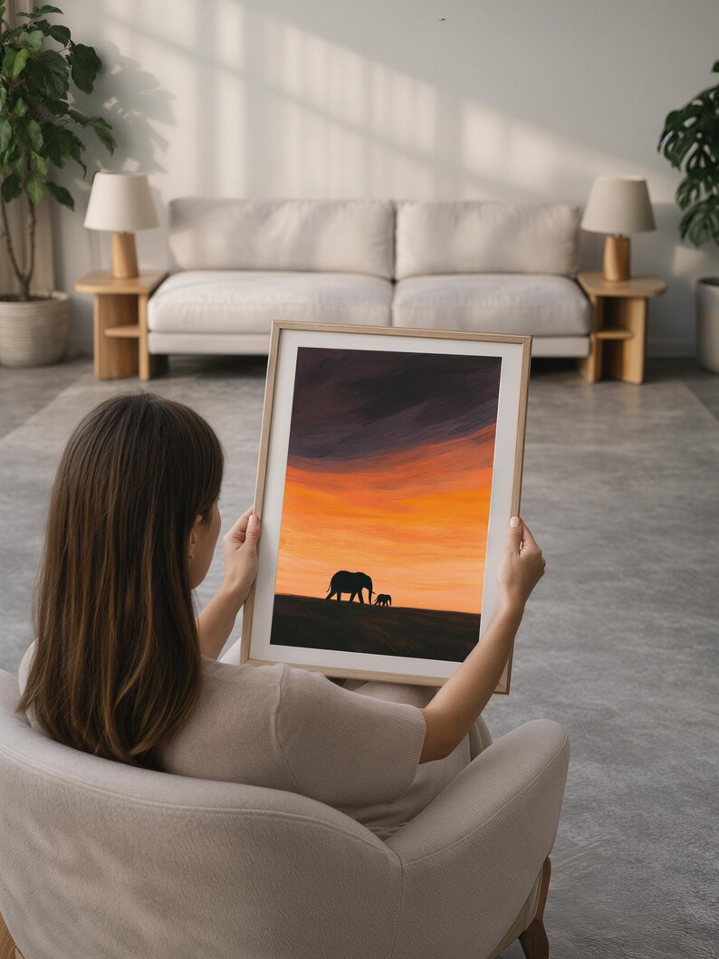May include: A framed art print featuring a sunset scene with silhouettes of two elephants. The sky transitions from dark purple to vibrant orange and yellow. The artwork is displayed in a light-colored wooden frame.