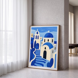 May include: A framed art print depicting a stylized village scene with white buildings, blue domes, and a blue staircase. The artwork is in a modern style, with bold colors and simple shapes. The frame is a light brown color.