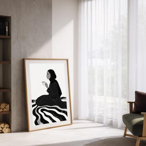 May include: A framed art print featuring a minimalist illustration of a person in a black garment holding a cup. The artwork is in black and white, with a striped pattern at the base. The frame is a light brown color.