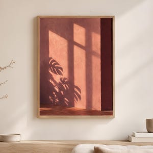 Puede incluir: Una impresión de arte mural rectangular en un marco de madera clara que presenta una fotografía de una pared de color terracota con sombras profundas proyectadas por un marco de ventana y la silueta de una hoja de planta monstera.