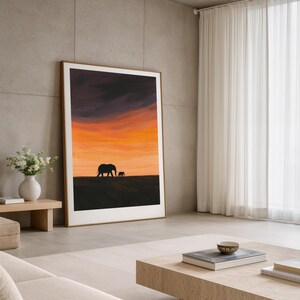 May include: A framed art print featuring a silhouette of two elephants against a vibrant sunset sky. The artwork has a warm color palette of orange, yellow, and purple. The print is displayed in a modern living room setting with neutral tones.