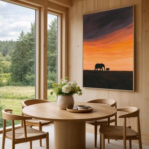 May include: A dining room with a round wooden table and four chairs. A framed artwork of an elephant silhouette against an orange and purple sunset hangs on the wall. A vase of white flowers sits on the table.