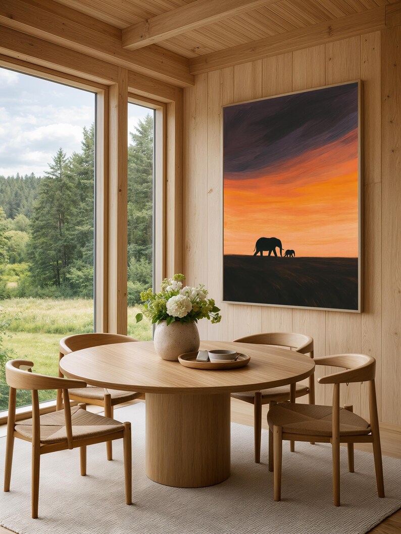 May include: A dining room with a round wooden table and four chairs. A framed artwork of an elephant silhouette against an orange and purple sunset hangs on the wall. A vase of white flowers sits on the table.