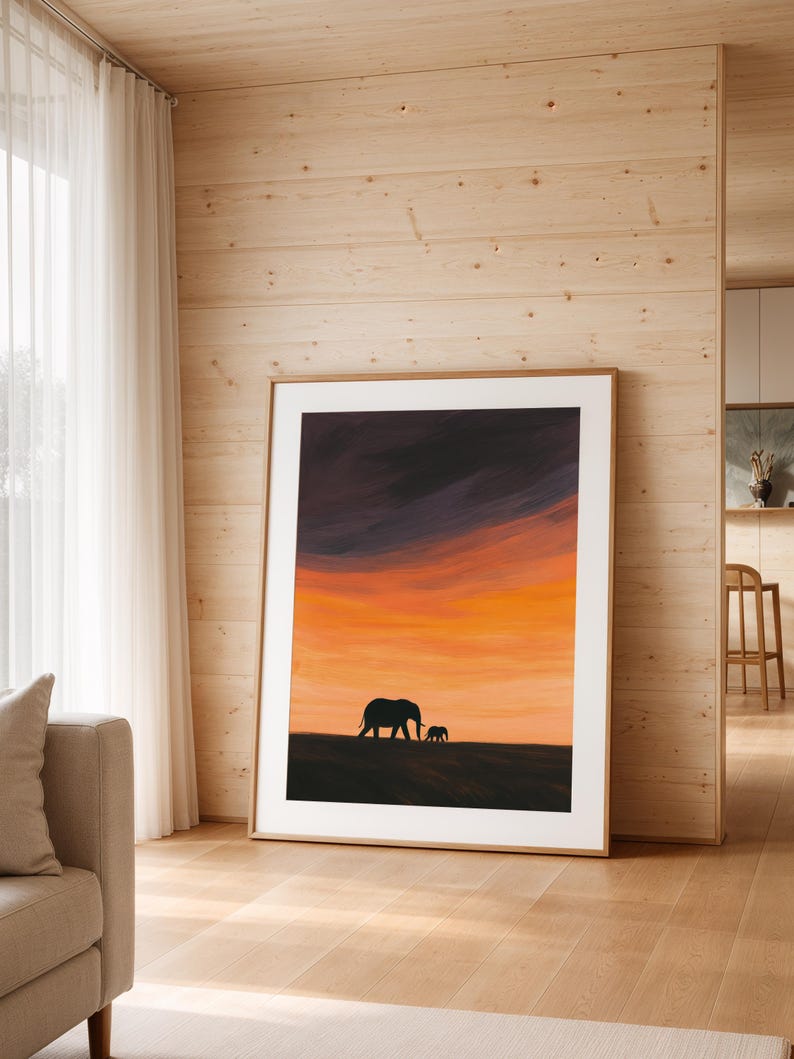 May include: A framed art print featuring a silhouette of two elephants against a vibrant sunset sky. The artwork showcases a warm palette of orange, yellow, and purple hues, creating a serene and naturalistic scene. The print is displayed in a light-colored wooden frame.