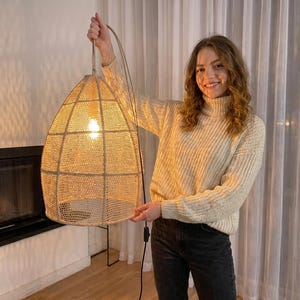 Puede incluir: Una lámpara colgante beige tejida con un diseño similar a una rejilla. La luz está suspendida por un cable beige y está iluminada por una sola bombilla. La lámpara está sostenida por una persona.