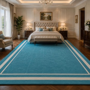 May include: A large rectangular area rug featuring a solid teal blue center field framed by a crisp white border. The rug has a flat, uniform texture and covers the floor in a room, providing a bold contrast to the surrounding neutral decor.