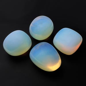 以下が含まれることがあります： 滑らかで丸みを帯びた形をした、研磨されたオパライトの石が4つ。石は半透明で乳白色の外観で、ソフトな青と黄色のグラデーションがあります。石は黒い背景に配置されています。