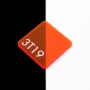 May include: A two-toned, diamond-shaped acrylic piece. One half is a deep brown, the other is bright orange. The number "3T19" is printed in white on the brown side. The edges are a vibrant pink. The background is split, with black on the left and white on the right.
