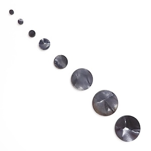 May include: An arrangement of eight dark gray, circular metal objects of varying sizes, arranged diagonally on a white background. The objects have a textured, slightly crumpled appearance, suggesting a hammered or forged finish. The overall effect is minimalist and modern.